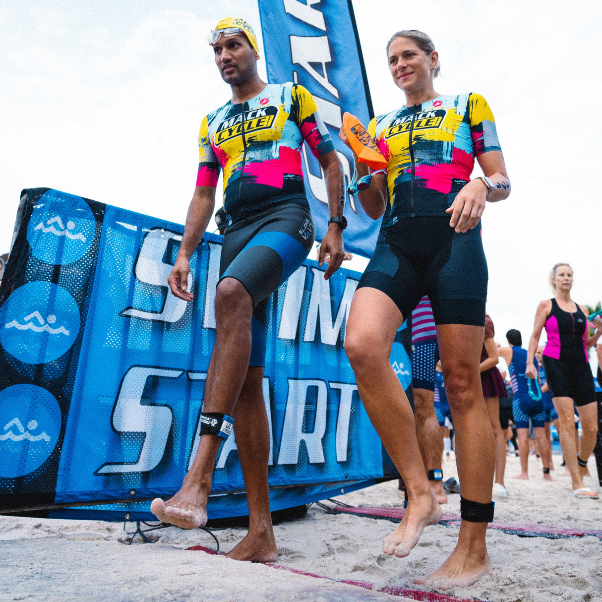 Two athletes in colorful Mack Cycle CMYK san remo custom tri suits. Walking on a beach during a triathlon with 'Start' branding in the background.