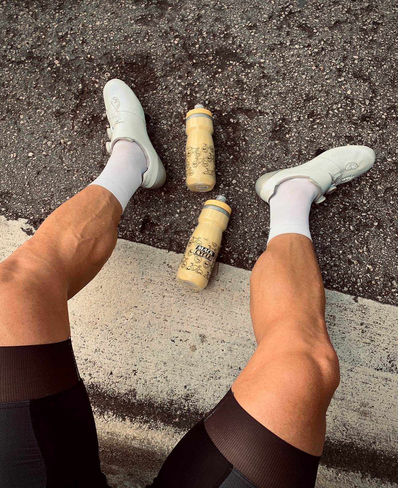 Man with strong legs wearing white Shimano S Phyre Shoes sitting on a curb with two yellow mack cycle Camelbak bottles