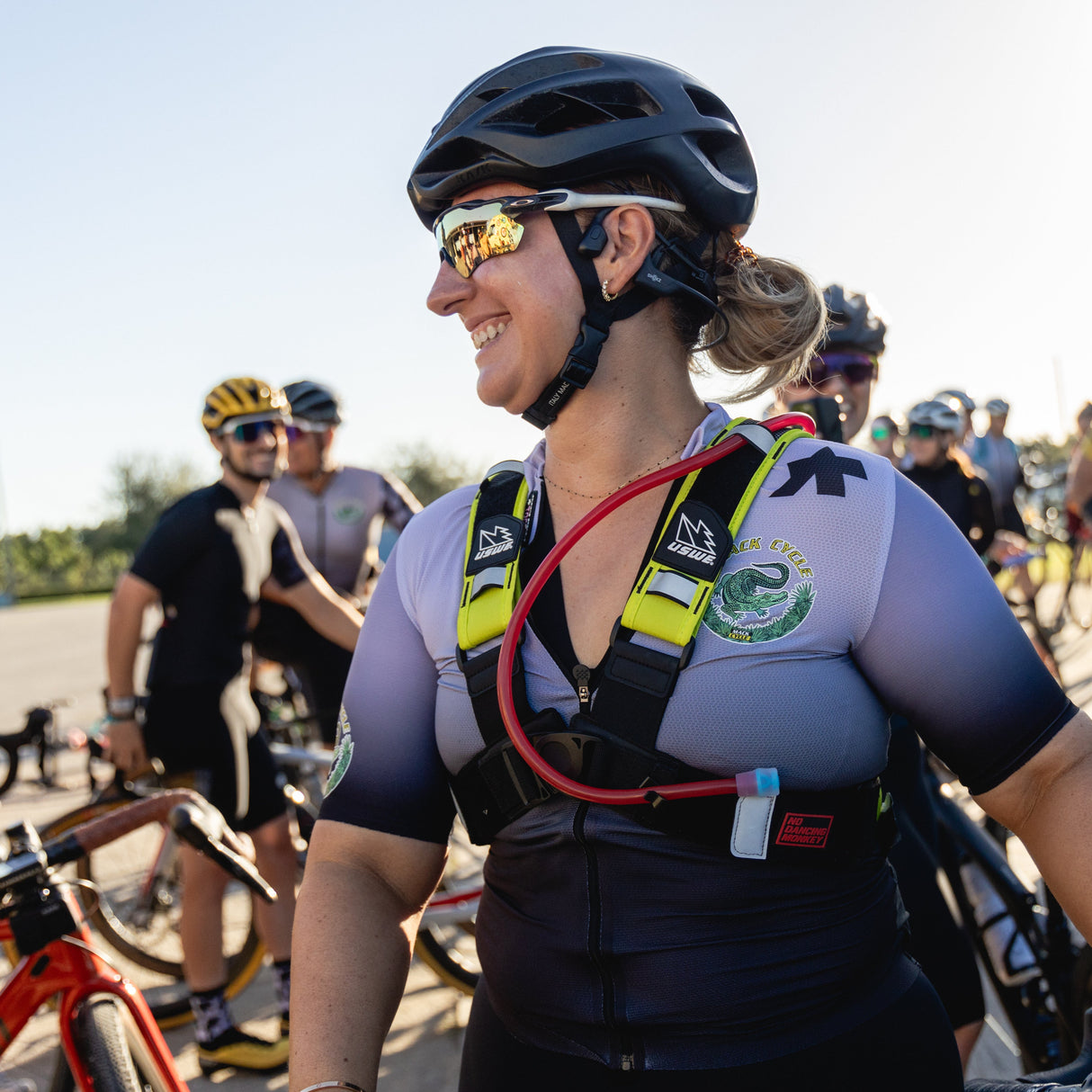 Woman in assos cycling gear wearing an USWE pack with a kask protone helmet and oakley sunglasses, standing among other cyclists at a gravel event.