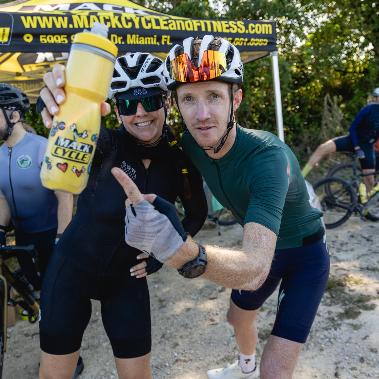 Maricarmen Saleta and Ian Boswell  posing with a yellow mack cycle brito camelbak water bottle under the mack cycle tent at the mack cycle gravel ride in miami.