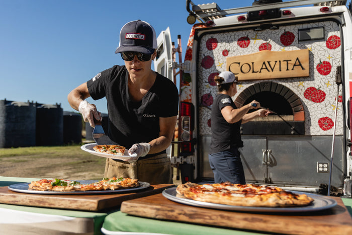 Mary Zider serving a hot fresh slice of pizza from the colavita pizza truck while ande sticks the next one into the brick oven to cook.