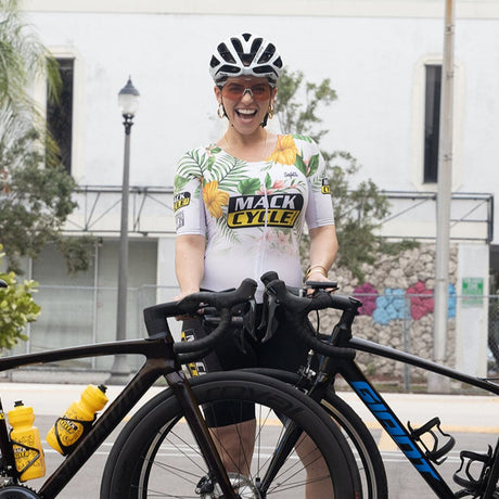 Janelle smiling with her new Specialized Tarmac SL7 outside of Mack Cycle. She is holding her new bike and her black and blue giant contend Trade in. She is wearing a white kask protons helmet and white floral tri suit. 