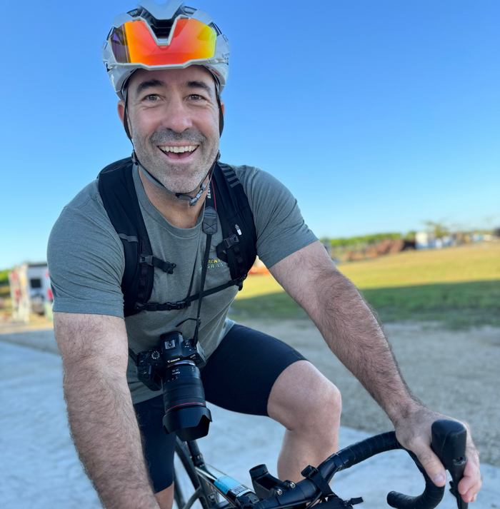 Man riding a gravel bike with a camera slung around his neck for the mack cycle gravel event